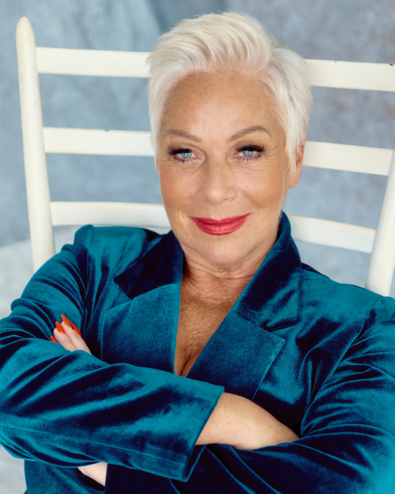 acting headshot of Denise Welch sitting on a white chair wearing blue velvety suit taken by photographer Kim Hardy taken in his London studio in front of a blueish background