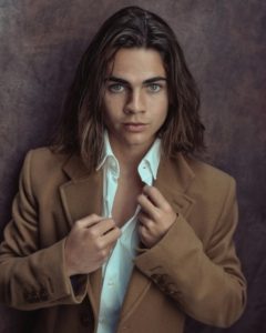 Headshot of young actor who appears in Wednesday taken by photographer Kim Hardy in his London studio in front of a burgundy backdrop wearing a brown jacket