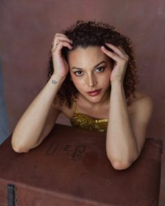 Acting headshot of young mixed race woman wearing a gold dress, leaning on her hands taken by photographer Kim Hardy in his London studio using a prop table and warm coloured background