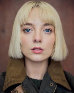 headshot of actor Kat Ronney featuring a woman with short blond hair, blue eyes in front of a warm coloured background taken by photographer Kim Hardy in his London studio