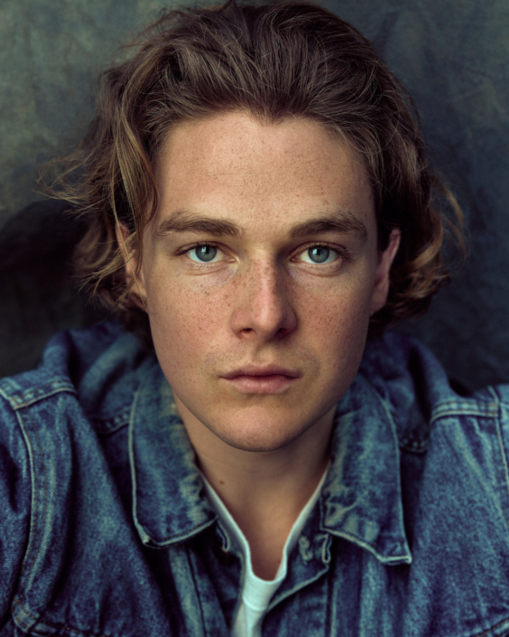 acting headshot of a young actor with blue eyes wearing a blue jeans jacket taken by Kim Hardy in his London studio in front of a hand-painted background