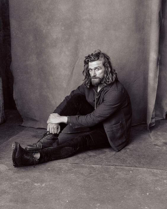 black and white headshot of actor Paul Bullion sitting in front of a hand-painted background in the London studio of Kim Hardy